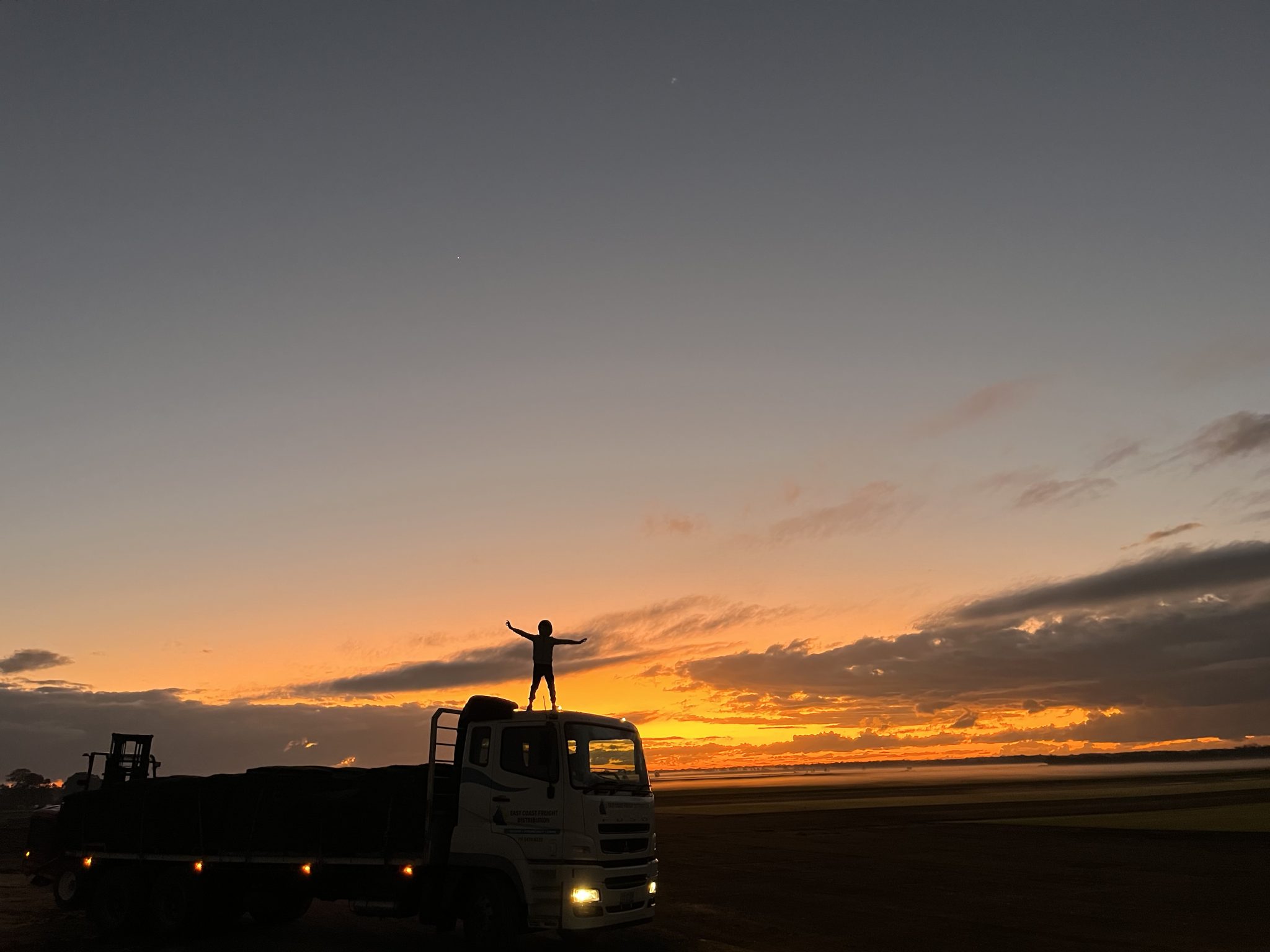 Silhouette of a young person standing on a truck cab with arms outstretched against a dramatic orange and yellow sunset over a misty field.